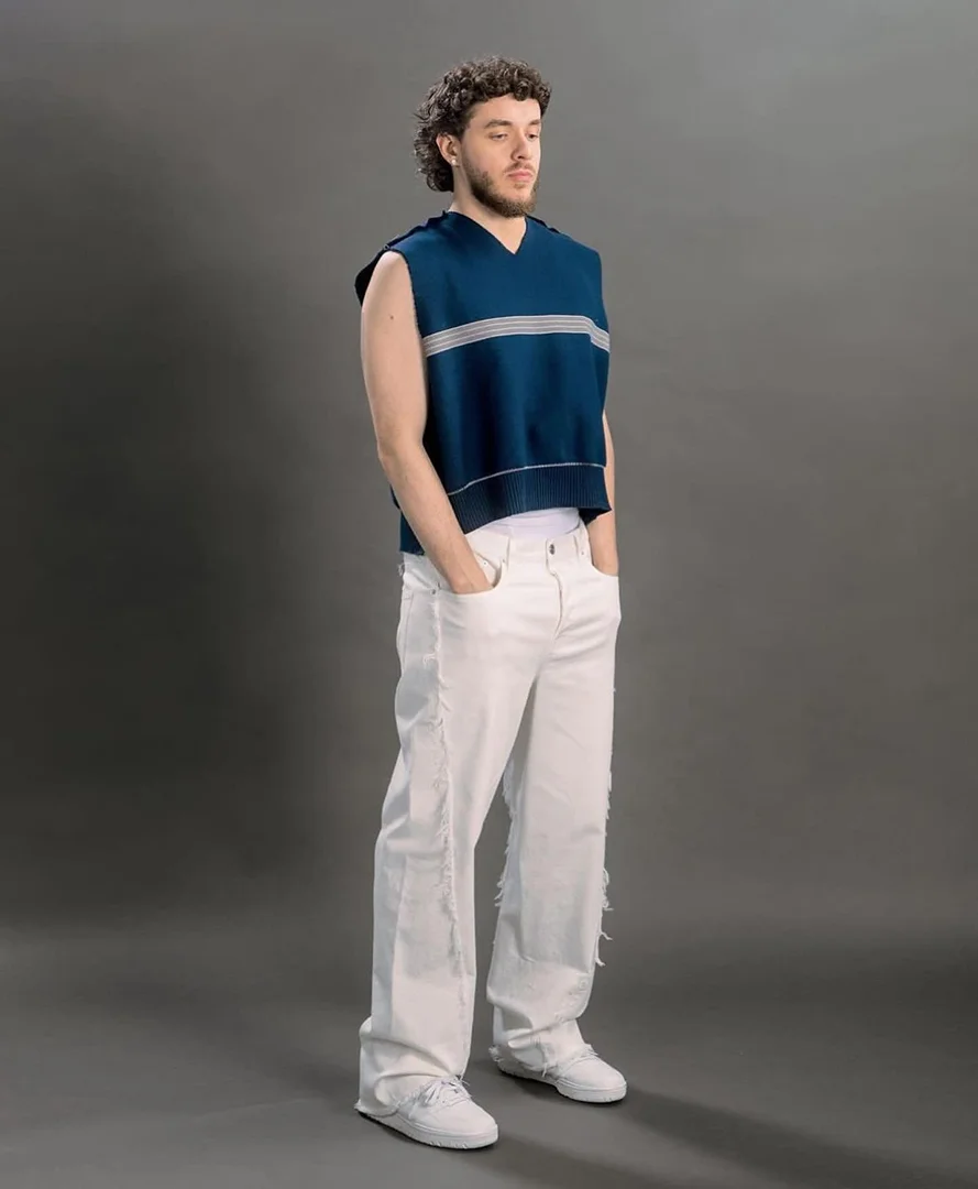 A full shot of musician Jack Harlow in white pants and a blue sweater vest standing against a grey background in a studio.