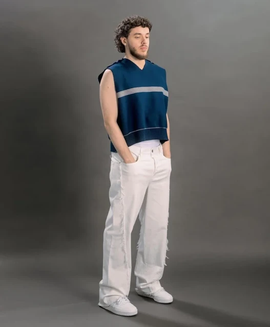 A full shot of musician Jack Harlow in white pants and a blue sweater vest standing against a grey background in a studio.