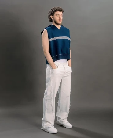 A full shot of musician Jack Harlow in white pants and a blue sweater vest standing against a grey background in a studio.