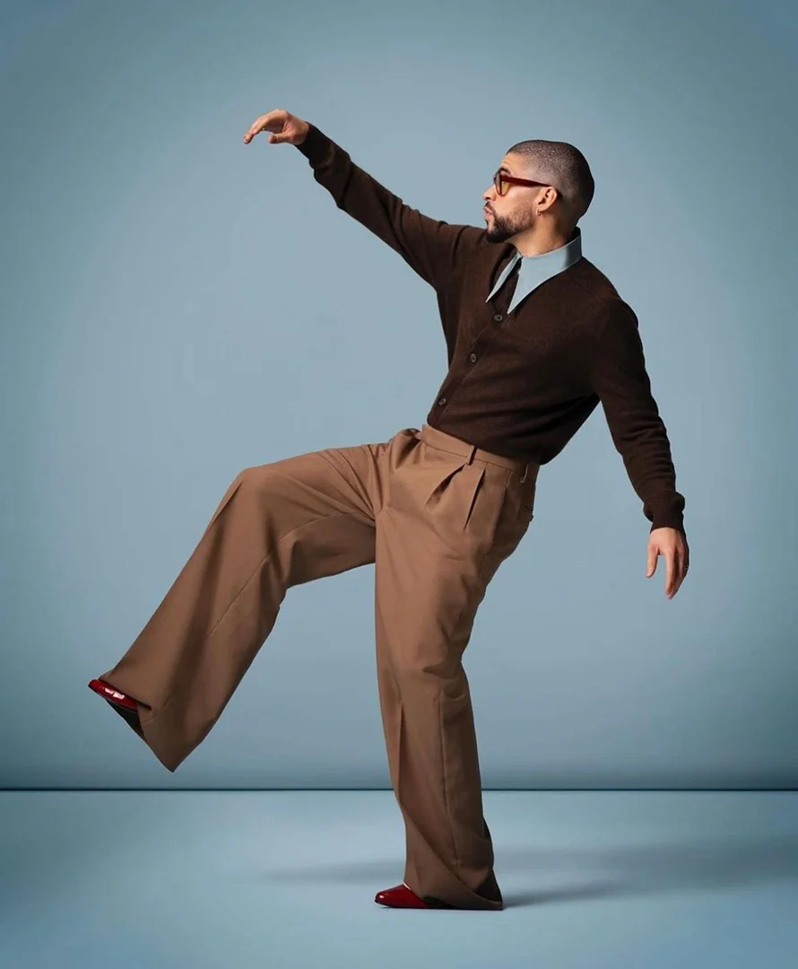 An adult man in a brown cardigan and wide-leg pants strikes a dynamic pose against a blue background, looking to the right.
