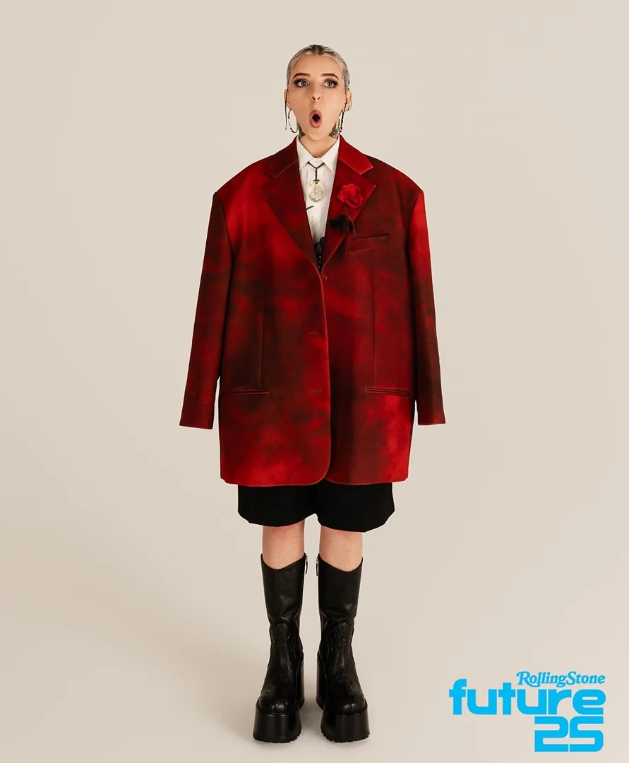 Full-length portrait of a young woman with blonde hair wearing a red blazer, shorts, and platform boots against a neutral backdrop.