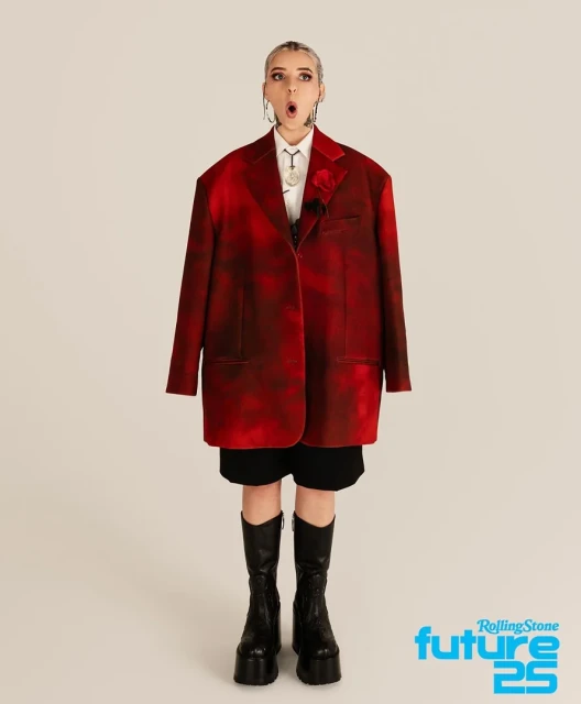 Full-length portrait of a young woman with blonde hair wearing a red blazer, shorts, and platform boots against a neutral backdrop.