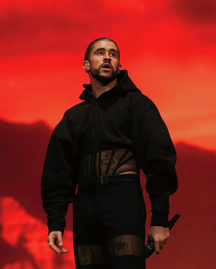 Bad Bunny performs on stage in a black hooded jacket with sheer detailing, black pants and boots against a red gradient background.