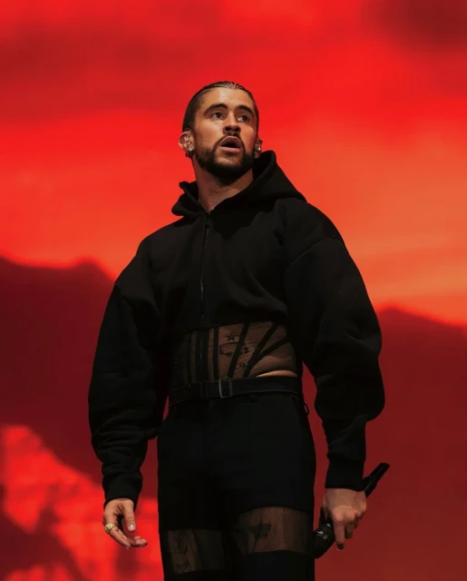 Bad Bunny performs on stage in a black hooded jacket with sheer detailing, black pants and boots against a red gradient background.