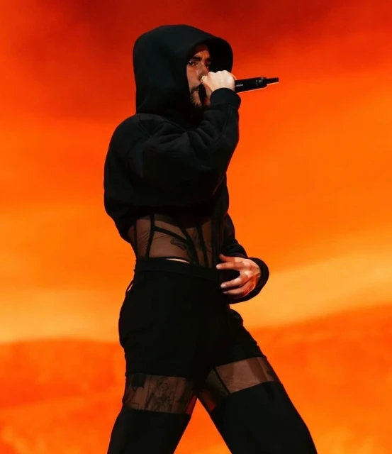 A male performer in a black hooded outfit and sheer corset is singing into a microphone on stage with a warm orange background.