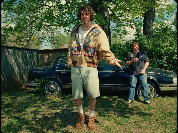 Two men pose in a grassy yard beside a navy car; one man wears shorts and the other jeans in outdoor, natural lighting.