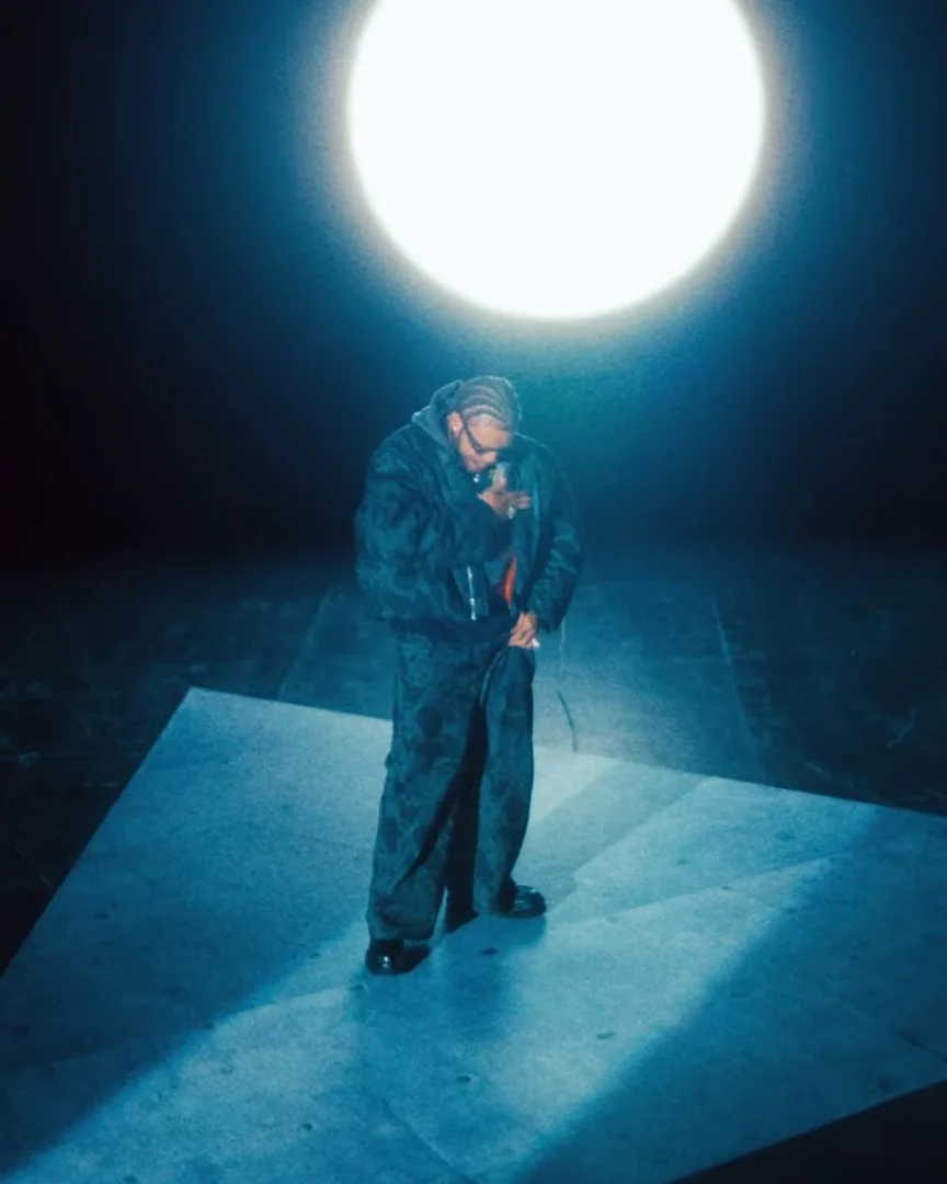 Adult male poses in a dark, moody atmosphere under a bright light, wearing a dark bomber jacket, matching pants, and black shoes.