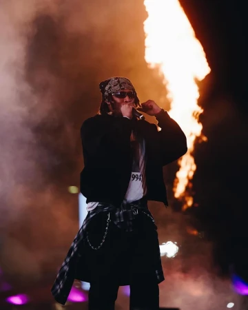 A musician sings into a microphone in front of flames on stage, wearing sunglasses and a bandana, with a flannel tied around his waist.