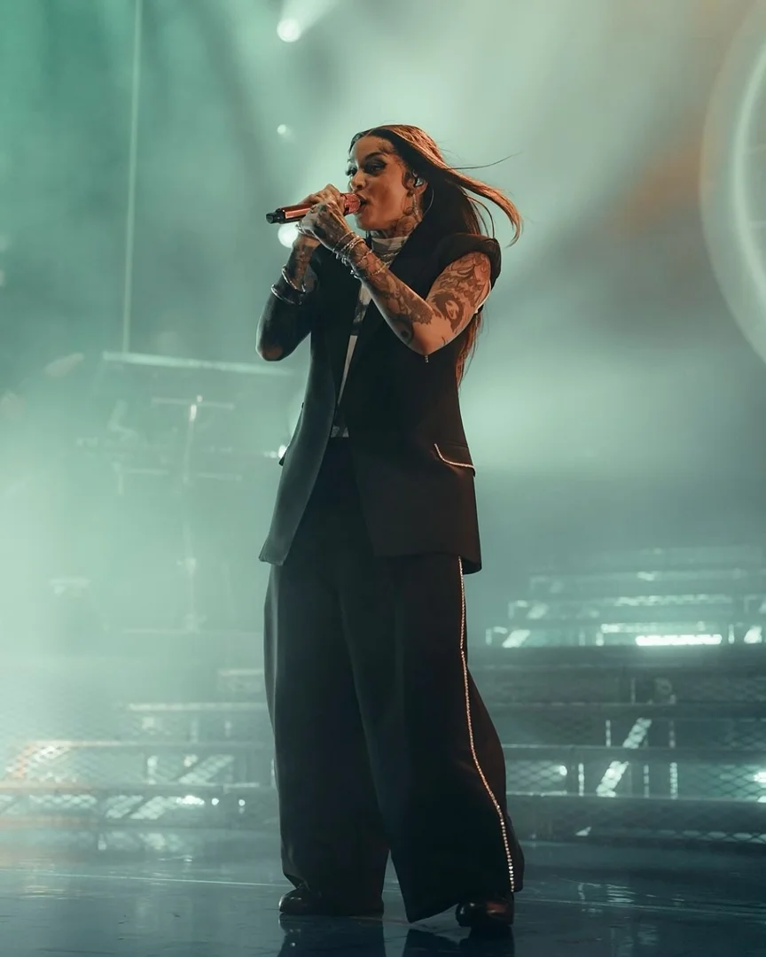A singer performs on stage in a black pantsuit, holding a microphone and singing in a well-lit venue with a stairway in the background.