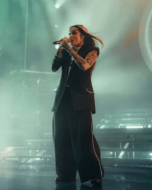 A singer performs on stage in a black pantsuit, holding a microphone and singing in a well-lit venue with a stairway in the background.