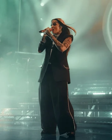 A singer performs on stage in a black pantsuit, holding a microphone and singing in a well-lit venue with a stairway in the background.