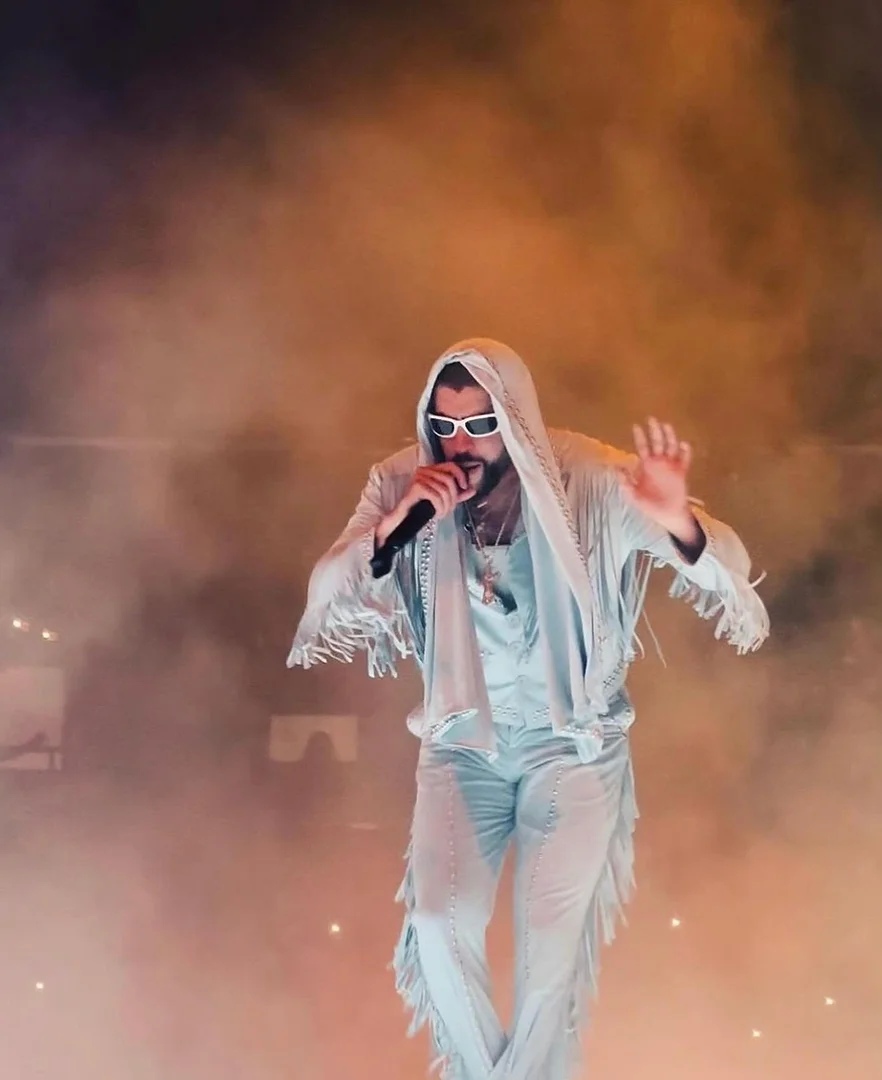 Bad Bunny performing on stage with a microphone, wearing sunglasses, a white fringed suit, and standing in front of a smoky background.