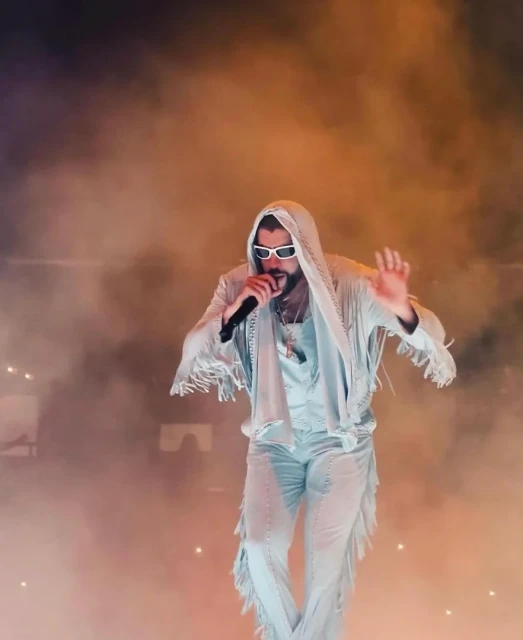 Bad Bunny performing on stage with a microphone, wearing sunglasses, a white fringed suit, and standing in front of a smoky background.
