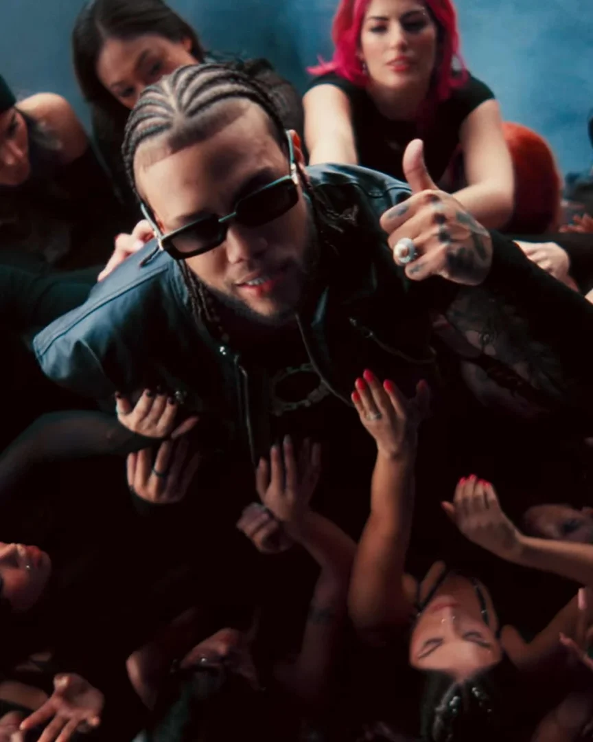 Man with sunglasses, braids, and a leather jacket gives a thumbs-up surrounded by women reaching toward him in a darkly lit scene.
