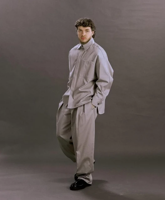 Full-length studio portrait of a young adult male with curly brown hair wearing a gray collared shirt and pleated gray pants.