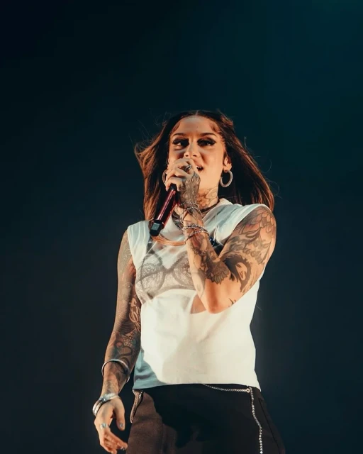 Singer Kehlani performing on stage with a microphone. She has tattoos on her arms and is wearing a white shirt and black pants.