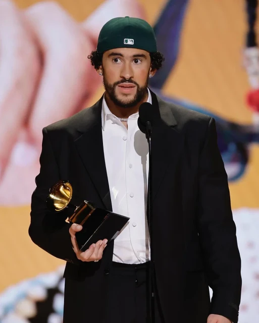 Bad Bunny accepts an award at a music event, wearing a black suit and a baseball cap while holding a trophy under bright lighting.