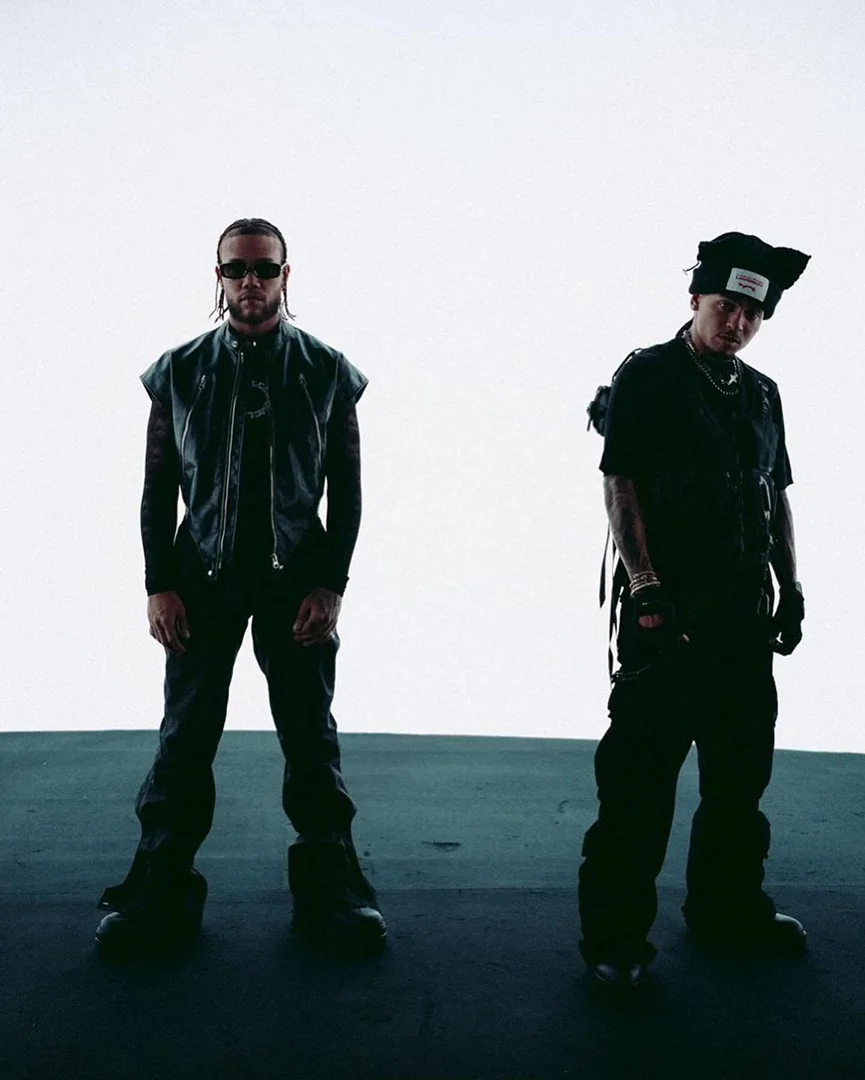 Two adult men stand in an indoor studio setting, wearing dark street fashion with tactical vests and bulky boots, under bright studio lighting.