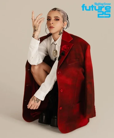 Portrait of a tattooed adult woman in a white shirt and red jacket squatting against a neutral background, looking at the camera.