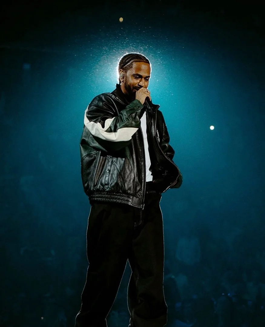 Adult male hip-hop artist performing on a stage with dramatic lighting in front of an audience. Wearing a leather jacket.