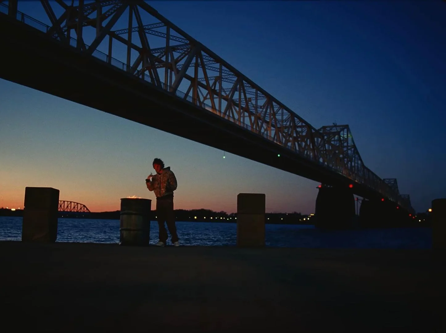 Man near barrel fire under a bridge at twilight on a city waterfront. River, sky, and city lights glow in the background.