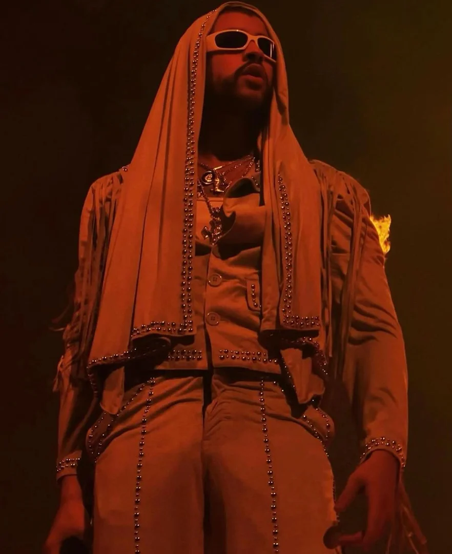 Bad Bunny performing on stage in an orange-toned, medium shot. He wears sunglasses and a hooded jacket with studded detail.