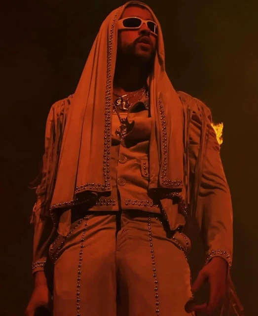 Bad Bunny performing on stage in an orange-toned, medium shot. He wears sunglasses and a hooded jacket with studded detail.