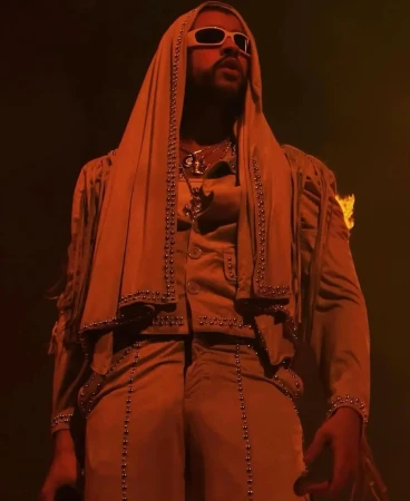 Bad Bunny performing on stage in an orange-toned, medium shot. He wears sunglasses and a hooded jacket with studded detail.