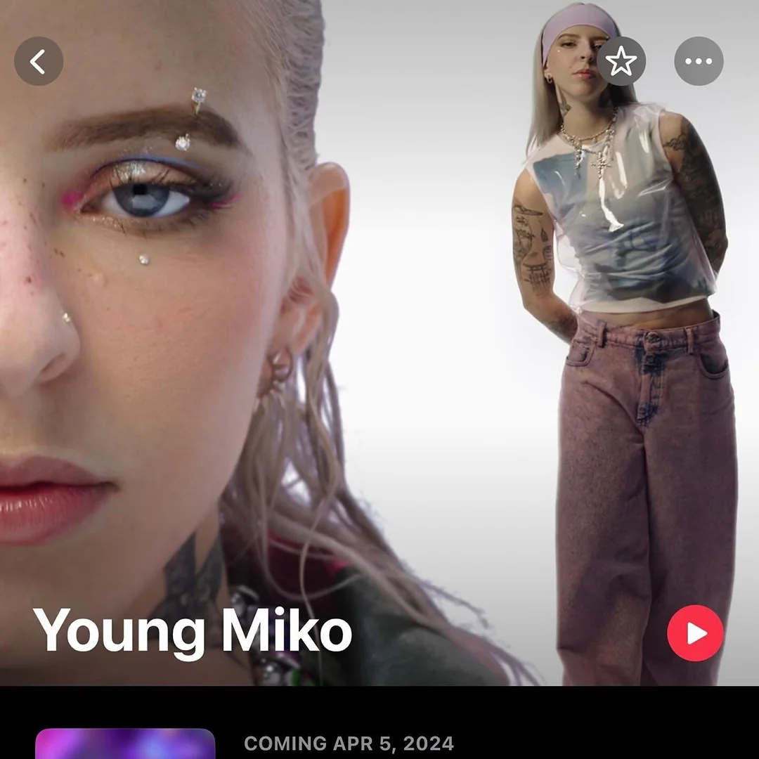 Blonde woman in pink pants and a sheer top with rhinestones on her face posing in front of a plain, light background in a studio.