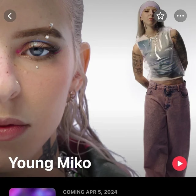 Blonde woman in pink pants and a sheer top with rhinestones on her face posing in front of a plain, light background in a studio.