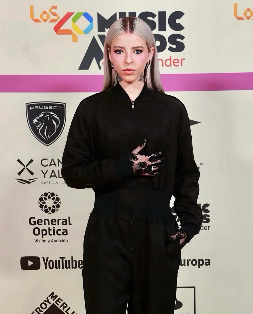 Singer poses on red carpet wearing a black tracksuit and lace gloves at Los 40 Music Awards event with logos and brands visible.