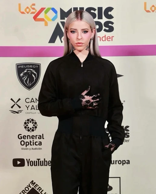 Singer poses on red carpet wearing a black tracksuit and lace gloves at Los 40 Music Awards event with logos and brands visible.