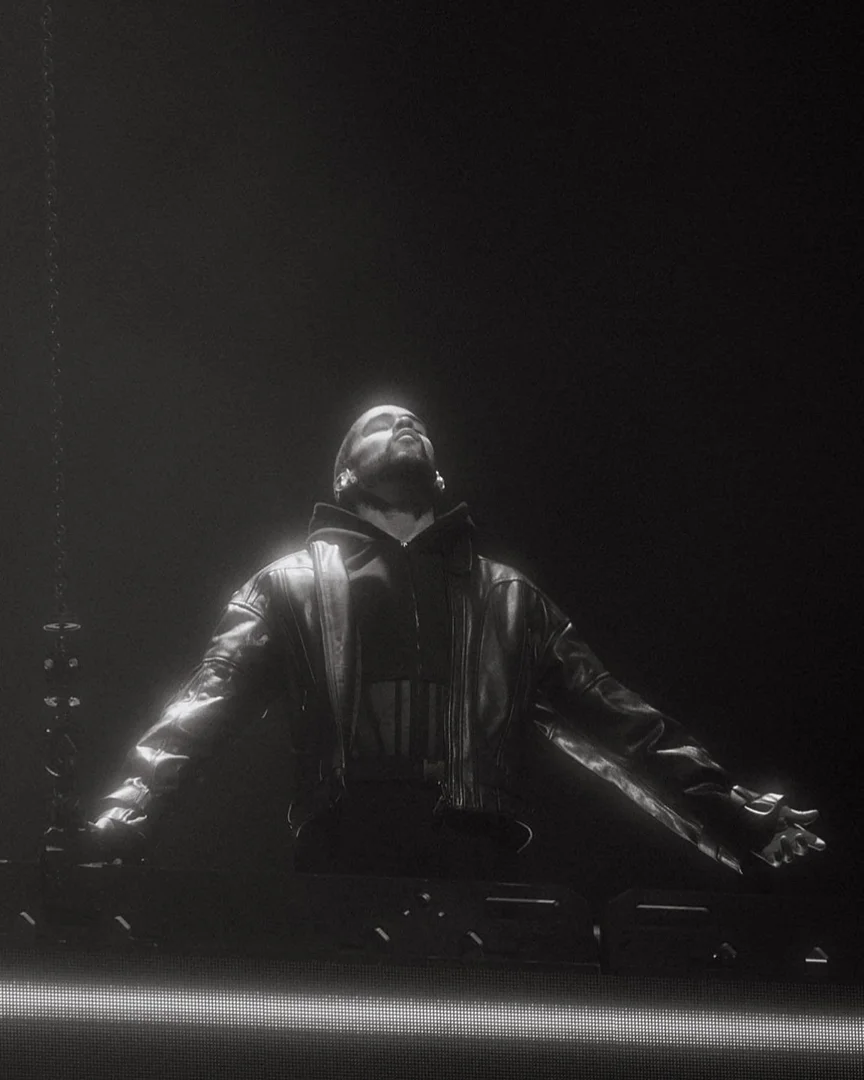 Man wearing a dark leather jacket and hooded sweatshirt stands with arms outstretched under dramatic spotlight, styled by Storm Pablo.