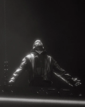 Man wearing a dark leather jacket and hooded sweatshirt stands with arms outstretched under dramatic spotlight, styled by Storm Pablo.