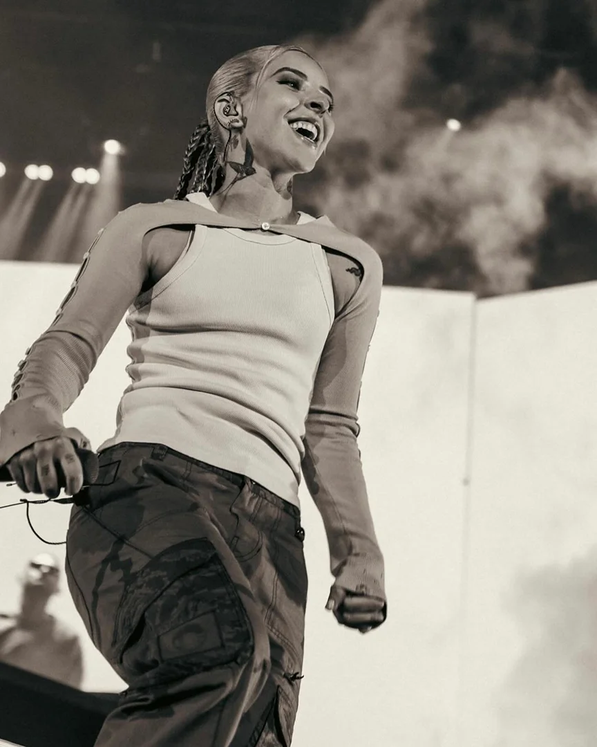 Monochromatic stage shot of a smiling woman with a visible neck tattoo, wearing camo pants and a tank top, performing with stage lights.