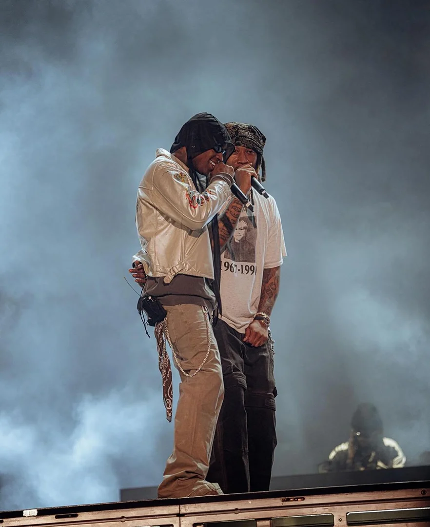 Two male performers sing on stage, wearing streetwear, bandanas, and microphones under hazy lighting, with a partial crowd in the background.