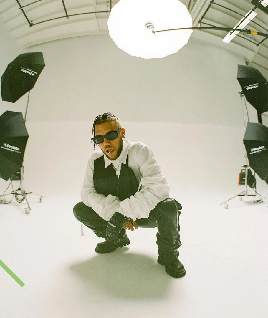 Man in a black vest and sunglasses squats in a white photo studio with lighting equipment. He is wearing black pants and boots.