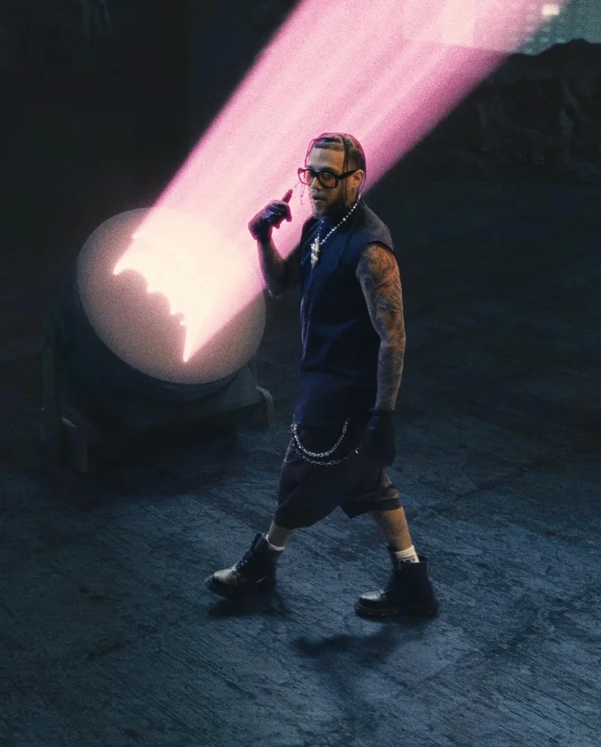 Man with tattoos wears glasses and a sleeveless top, walking toward bright pink stage lights in a dimly lit interior setting.