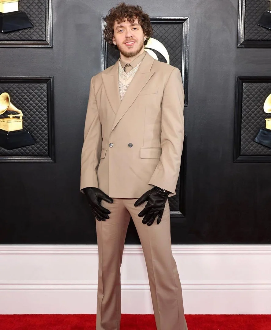Jack Harlow poses on a red carpet in a tan suit, gloves, and patterned shirt, surrounded by rows of Grammy Award plaques.