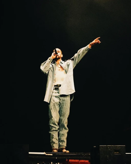 Male music artist performing on stage with a microphone. He is wearing a light-wash denim outfit and pointing to the right with his hand.