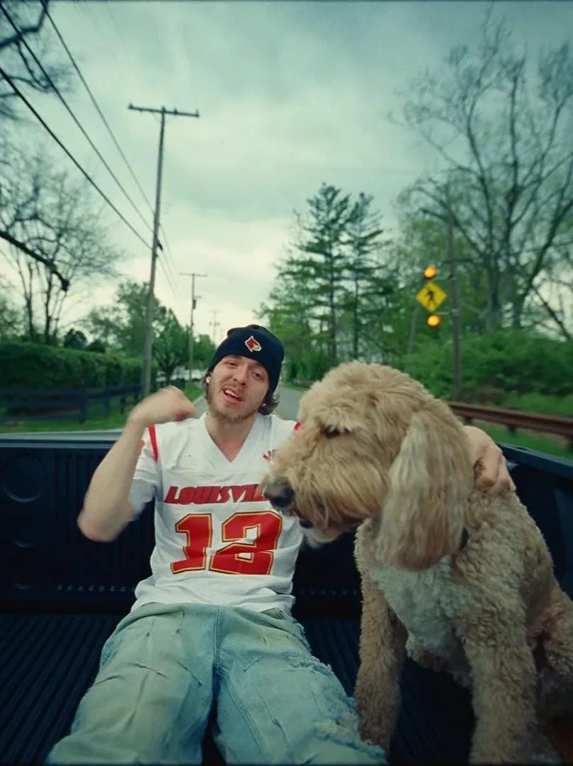 Man and Goldendoodle dog in the back of a truck outdoors, wearing a sports jersey and hat on a cloudy day.