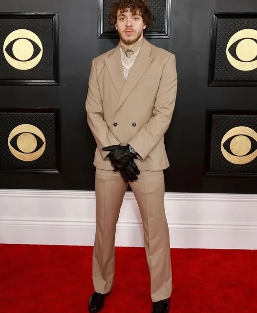 Jack Harlow in a tan suit poses on a red carpet with black Gramophone designs on the wall during an award show event.