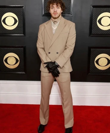 Jack Harlow in a tan suit poses on a red carpet with black Gramophone designs on the wall during an award show event.