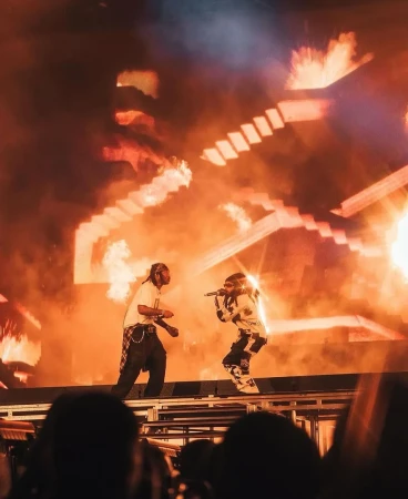 Two musicians perform on stage with pyrotechnics in the background, wearing streetwear fashion and singing into microphones during concert.
