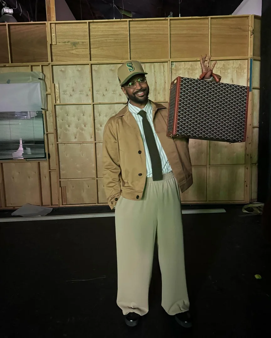 Stylish man poses in a tan jacket, light pants, and baseball cap while holding a patterned briefcase against a wood-paneled wall.