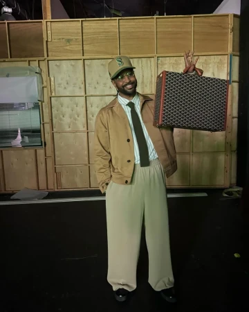 Stylish man poses in a tan jacket, light pants, and baseball cap while holding a patterned briefcase against a wood-paneled wall.