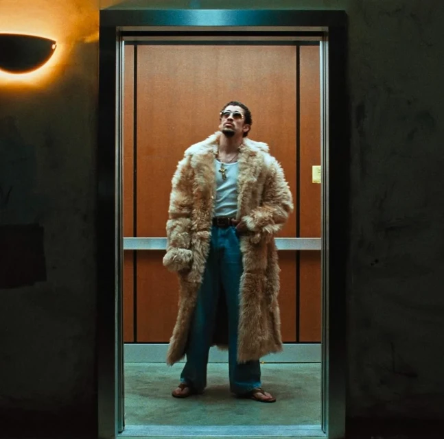 Adult man wearing fur coat and sunglasses in elevator, looking up. He has a goatee and is wearing a gold chain and open-necked shirt.