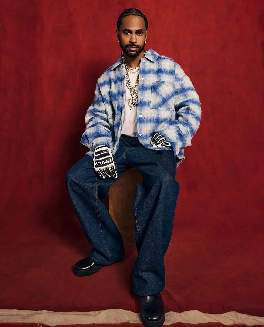 A full-length portrait of a man wearing a plaid shirt, jeans, and gloves, seated against a red backdrop in a studio environment.