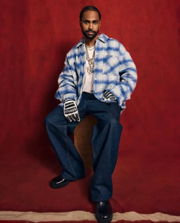 A full-length portrait of a man wearing a plaid shirt, jeans, and gloves, seated against a red backdrop in a studio environment.