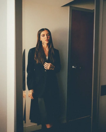 Adult female in a black coat stands in front of a mirror in an indoor environment with neutral and dark color tones.